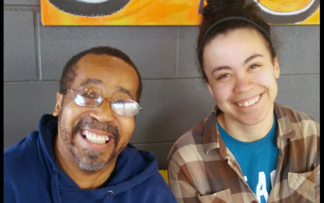 Man with glasses and blue hooded sweatshirt sits next to young woman with headband in hair and plaid shirt