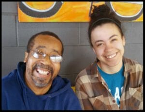Man with glasses and blue hooded sweatshirt sits next to young woman with headband in hair and plaid shirt