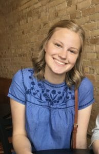 Young woman with long blond hair and blue shirt with flower embroidery smiles in front on brick wall