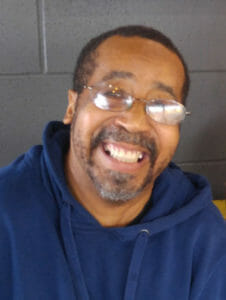 Black man with glasses and short mustache and beard wearing blue hooded sweatshirt smiles at camera