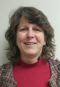 Smiling woman with wavy hair wearing red mock turtleneck and tweed blazer