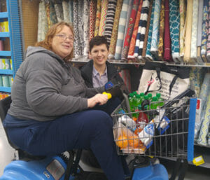 Woman driving motorized cart filled with items and another young woman smile in front of display of fabric bolts