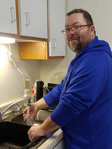 Man with glasses and beard holds hand under running water at kitchen sink