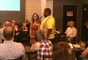 Man in yellow shirt and orange safety suspenders holds a microphone while a crowd looks on