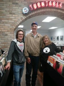 Tall man in tan jacket stands between young man and middle-age woman in record store under neon Turntable sign
