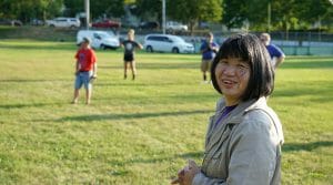 Woman in tan jacket smiles at camera with four people in the outfield on a softball field behind her