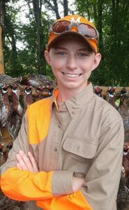 Teenage boy in tan and blaze orange shirt stands in front of hanging pheasants