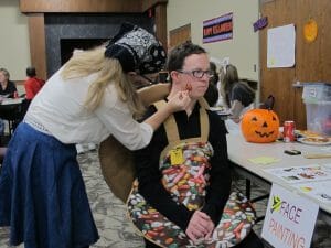 Young man wearing a candy bowl costume sits at a face painting station while a teenage girl paints on his cheek